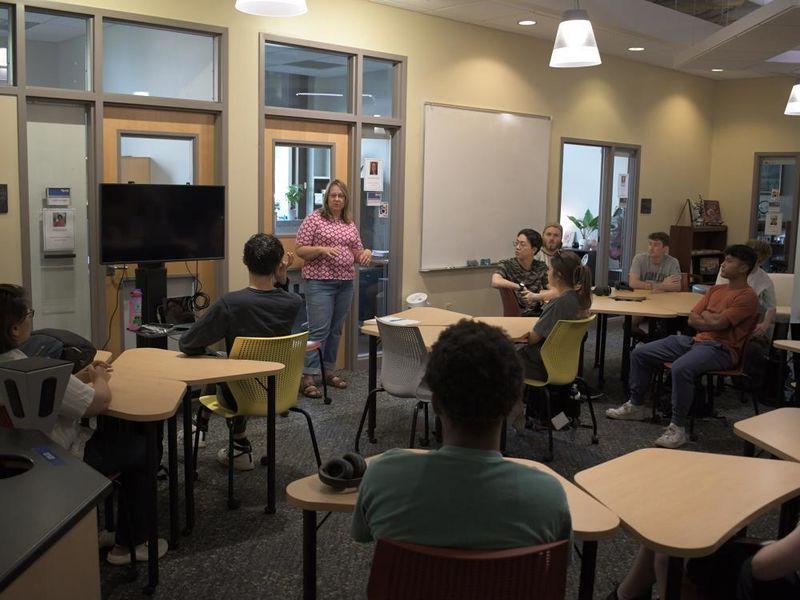 Person stands at the front of a room, talking to other people sitting at tables. 